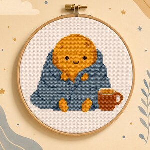 May include: A cross-stitch embroidery in a round wooden hoop featuring a yellow, rounded character wrapped in a blue blanket. The character has a simple face with two black eyes and a small smile. A brown mug sits next to the character on white fabric.