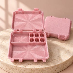 May include: A pair of rectangular, shimmering pink plastic storage cases. One case is open, revealing a divided interior with a large rectangular compartment and six circular wells. The other case is closed, showing a textured, glittery surface.