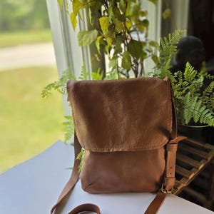 May include: A brown leather crossbody bag featuring a fold-over flap, a long adjustable shoulder strap, and a side buckle detail. The bag has a textured surface and a rectangular shape with rounded bottom corners, providing a simple design.