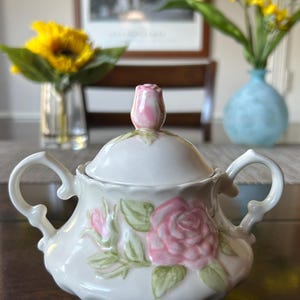 May include: A white ceramic sugar bowl with two curved handles and a matching lid topped with a pink rosebud finial. The body of the bowl features a raised, hand-painted design of a large pink rose with green leaves and a smaller rosebud.