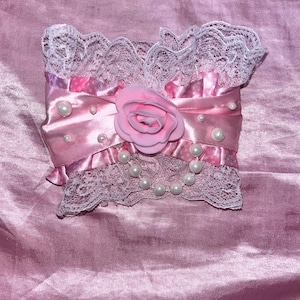 May include: A pink satin and white lace cuff featuring a central pink rose applique, scattered white faux pearls, and a draped string of pearls. The accessory is constructed with ruffled white lace trim at the top and bottom edges.
