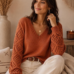 May include: A rust-colored V-neck sweater featuring long sleeves with an intricate open-knit diamond pattern and ribbed cuffs. The sweater is paired with off-white trousers and a brown leather belt with a gold-toned buckle.