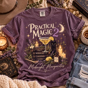 May include: A purple short-sleeve t-shirt featuring a graphic print with the text Practical Magic 2 and Midnight Margaritas. The design includes a black cat, a margarita glass, stacked books labeled Salt, Rosemary, Lavender, Love, and candles.