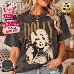 Può includere: Una t-shirt grigio antracite con effetto lavaggio acido, caratterizzata da una grande stampa grafica color marrone chiaro raffigurante una donna con capelli voluminosi e la scritta DOLLY in grandi lettere maiuscole sopra il ritratto. Maniche corte e girocollo.