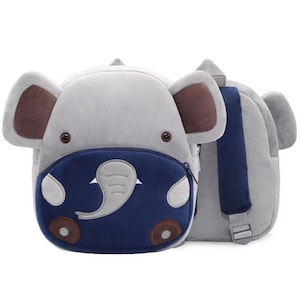 May include: A plush backpack shaped like an elephant with large gray ears, dark brown inner ear accents, and small dark eyes. The front features a dark blue zippered pocket with a light gray elephant trunk applique, white tusks, and brown feet.