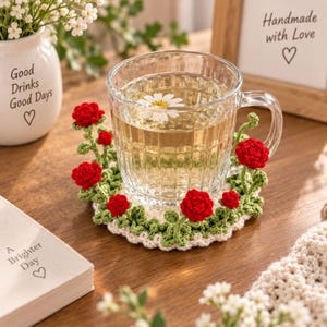 May include: A round crochet coaster featuring a white base with green leaves and small red roses, supporting a clear glass mug filled with tea and a daisy. The coaster is placed on a wooden surface near a white mug with the text Good Drinks Good Days.