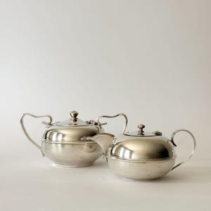 May include: A silver-toned metal sugar bowl with two curved handles and a matching lid, positioned next to a silver-toned metal teapot featuring a curved spout, a single handle, and a lid with a small knob finial on top of each piece.