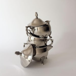 May include: A stack of three polished silver-toned metal serving vessels, including two lidded sugar bowls and one creamer. Each piece features ornate, detailed handles and feet with a floral motif, and the lids are topped with a finial.
