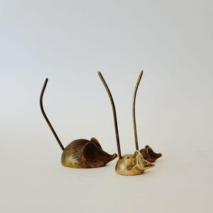 May include: Three brass mouse figurines of varying sizes, each featuring a smooth, rounded body, pointed ears, and a long, thin, upright metal tail. The metallic surfaces show a natural patina with subtle variations in golden and bronze tones.
