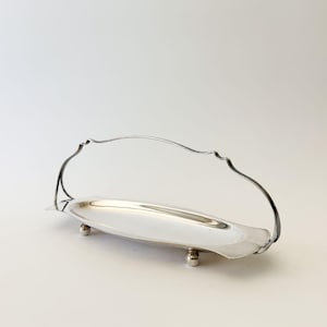 May include: A silver-toned metal serving tray with an elongated oval shape, featuring a high, arched wire handle and four small, rounded ball feet. The tray has a smooth, reflective surface and a simple, clean design for holding small items.