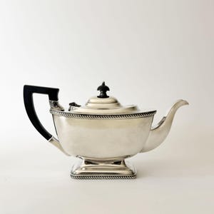 May include: A silver-toned teapot featuring a polished metallic body with a rectangular base and a rope-style border detail. It includes a black handle and a matching black finial on the lid, paired with a curved spout extending from the side.