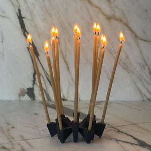 May include: A black multi-candle holder with a star-shaped base holding ten thin, light-coloured taper candles. Each candle is lit, producing a small, bright orange flame. The holder is constructed with individual slots for each candle.