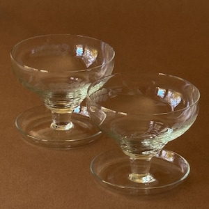 May include: Two clear glass coupes with wide, shallow bowls and short, thick stems resting on circular bases. The glass features delicate, etched floral patterns along the rim of the bowls, providing a subtle decorative detail to the glassware.