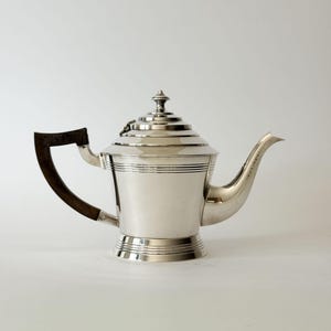 May include: A silver-coloured teapot featuring a stepped, conical lid with a small finial, a curved spout, and a dark brown, angular handle. The body of the teapot is smooth and metallic with horizontal grooved bands around the base and neck.