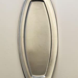 May include: A long, narrow, oval-shaped serving tray made of polished silver-toned metal. The tray features a smooth, recessed centre area with a wide, slightly raised rim that follows the elongated, rounded contour of the overall piece.