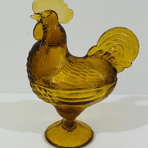 May include: An amber-colored glass rooster-shaped candy dish with a lid. The piece features a detailed molded texture representing feathers on the body and tail, a prominent comb on the head, and a circular pedestal base for stability.