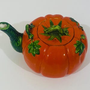 May include: A ceramic teapot shaped like a bright orange pumpkin with a green stem handle on the lid. The spout is textured like green leaves, and the body features embossed pumpkin ridges with small green leaf accents on the sides.
