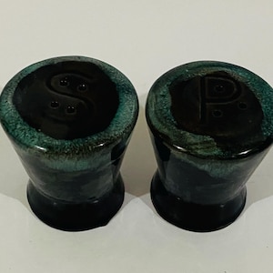 May include: A pair of ceramic salt and pepper shakers with a glossy finish. The shakers feature a dark black base that transitions into a vibrant teal rim. The top of each shaker is marked with a letter, S for salt and P for pepper, with holes.