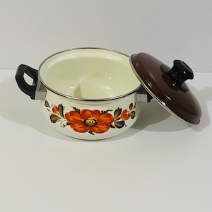 Peut inclure: Une marmite en émail de couleur crème avec un motif floral composé de fleurs orange et marron sur l'extérieur. La marmite possède deux poignées latérales noires et un couvercle marron assorti avec un bouton noir. L'intérieur est crème.