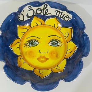 Könnte beinhalten: Eine runde Keramikschale mit gewelltem Rand, die eine handbemalte gelbe Sonne mit Gesicht, blauen Augen und roten Wangen zeigt. Der Hintergrund ist dunkelblau und der Text O' Sole mio steht in schwarzer Schrift oben im Schaleninneren.