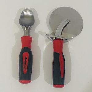 May include: A metal ice cream scoop and a circular pizza cutter, both featuring ergonomic black and red handles with the Snap-on brand logo printed in white on the scoop handle. The tools have polished silver-coloured metal heads and components.