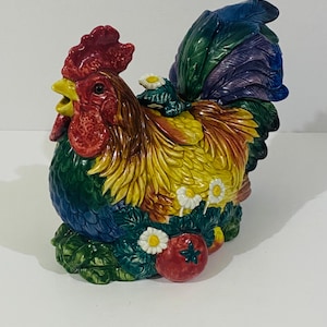 Może przedstawiać: Ceramiczny dzbanek w kształcie koguta z żywą, wielokolorową glazurą. Kogut ma czerwony grzebień i dzwonki, żółte pióra na szyi oraz niebiesko-zielony ogon. Podstawa ozdobiona jest rzeźbionymi zielonymi liśćmi, białymi stokrotkami i czerwonym pomidorem.