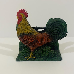 May include: A cast metal cockerel figurine featuring a vibrant red comb and wattle, a yellow neck, and a brown body. The cockerel stands on a textured green base that resembles grass, with a black fence-like structure positioned behind the bird.