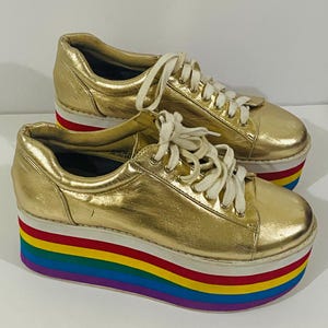Può includere: Un paio di sneakers color oro metallizzato con lacci bianchi e una spessa suola a piattaforma. La suola a piattaforma è decorata con strisce orizzontali nei colori rosso, giallo, verde e blu. Le sneakers hanno una punta arrotondata.