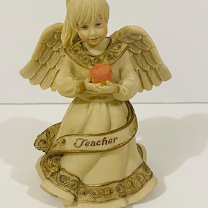 May include: A cream-colored angel figurine with feathered wings, wearing a long gown with gold-trimmed edges. The angel has a ponytail, holds a small red apple in both hands, and features a banner across the front with the word Teacher printed.