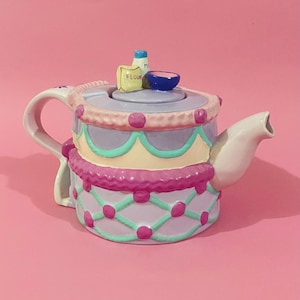May include: A ceramic teapot shaped like a tiered cake with a lavender base, pink lattice pattern, and teal accents. The lid features miniature sculpted items including a bag labelled FLOUR, a milk bottle, and a small blue bowl. It has a white handle and spout.