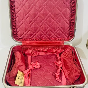 Peut inclure: Une valise grise ouverte avec un cadre en métal argenté et une doublure intérieure matelassée rouge foncé. L'intérieur présente des poches en tissu froncé sur les côtés, des sangles de maintien et une étiquette beige avec le texte KEY.
