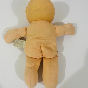 Peut inclure: Une poupée couleur pêche avec une tête ronde et chauve et un corps recouvert de tissu doux. La poupée présente un petit détail cousu en forme de queue sur le bas du dos et une étiquette en tissu blanc attachée sur le côté avec du texte imprimé.