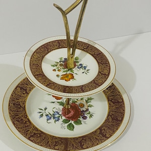 May include: A two-tiered serving stand featuring two round white porcelain plates with a deep red and gold filigree border. Each plate is decorated with a central floral bouquet. A gold-toned metal handle connects the two tiers vertically.