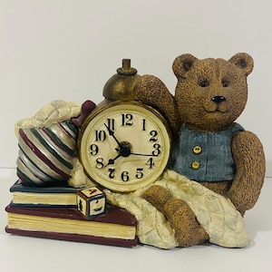 May include: A brown teddy bear figurine wearing a blue vest, positioned next to a round analogue clock with a gold frame. The clock sits atop two stacked books, one dark blue and one maroon, alongside a small alphabet block and a striped fabric bag.