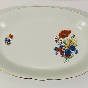 Op de afbeelding: Een ovale witte porseleinen serveerschaal met een geschulpte rand en een dunne gouden rand. De schaal is voorzien van twee bloemstukken met rode klaprozen, blauwe korenbloemen en gele tarwearen op het witte keramische oppervlak.