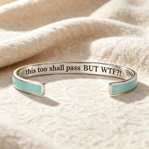 May include: A silver-toned cuff bracelet featuring a light blue enamel inlay along the outer band. The interior surface of the metal bracelet is engraved with the black text, "this too shall pass BUT WTF?!" in a clear, sans-serif font style.