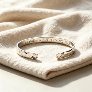 May include: A silver-toned metal cuff bracelet with a hammered, textured exterior surface. The inner band is engraved with the black text "This too shall pass, BUT FUCKING HELL" and features a small heart symbol near the open end of the bracelet.