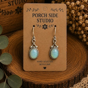 May include: A pair of silver-toned drop earrings featuring oval-shaped iridescent stones with a rope-textured border. The earrings hang from silver hooks and are displayed on a rectangular brown card labeled Porch Side Studio.