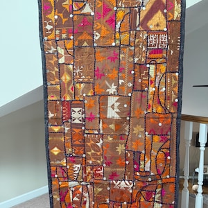 Op de afbeelding: Een rechthoekige wandhanger met een patchworkontwerp van bruine stoffen panelen. Het stuk is versierd met ingewikkeld borduurwerk in tinten oranje, roze en wit, geaccentueerd door kleine ronde spiegeltjes die over het hele oppervlak zijn gestikt.