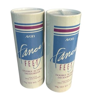 Könnte beinhalten: Zwei weiße, zylindrische Avon Fancy Feet Doppelwirkungs-Deodorierer für Füße, Schuhe und Sneaker. Jeder Behälter zeigt blaue und violette horizontale Streifen, das Avon-Logo und die Aufschrift 99g net.