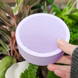 May include: A round, light purple portable speaker featuring a flat top surface with a dense grid of small, circular perforations. The device has a smooth, matte finish and a cylindrical body, presented in a compact, handheld size.