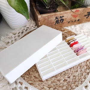 May include: A white rectangular plastic storage box with a textured lid and a divided interior tray. The tray contains several small cardboard bobbins wrapped with embroidery floss in shades of tan, brown, red, light pink, and purple.