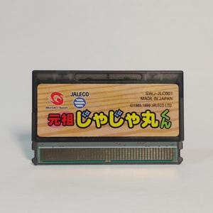 以下が含まれることがあります： WonderSwan用ゲームソフトの黒いプラスチック製カートリッジ。ラベルは明るい木目調のデザインで、JALECO、SWJ-JLC001、MADE IN JAPAN、1985, 1999 JALECO LTDという文字と、日本語のタイトル「元祖じゃじゃ丸くん」が印刷されています。