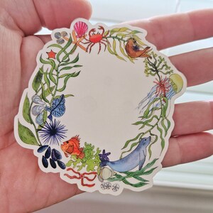 May include: A colorful sticker featuring a wreath of seaweed, sea creatures, and shells. The wreath is surrounded by a white background and has a blue seal in the center.