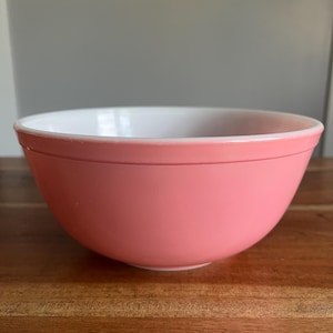 May include: A round mixing bowl with a smooth, solid pink exterior and a white interior. The bowl features a simple, wide-rimmed design with a slightly tapered base, constructed from a opaque, glass-like material with a glossy finish.