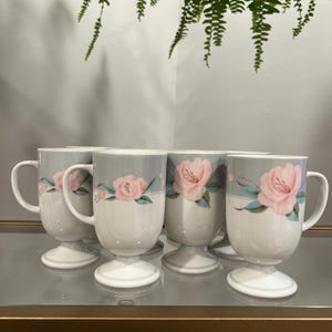 以下が含まれることがあります： ペデスタルベース付きの白いセラミック製マグカップ4個セット。上部にグレーの帯が施され、緑の葉を伴うピンクのバラの花柄が描かれています。マグカップには曲線的なハンドルが付いており、全体的に滑らかで光沢のある仕上がりになっています。