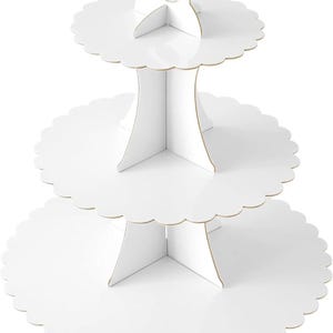 May include: A three-tiered white cupcake stand constructed from cardboard. The stand features three circular tiers with scalloped edges, supported by interlocking vertical panels that form a central column with a handle at the top.