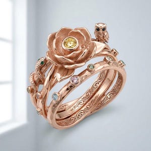 Puede incluir: Un anillo de oro rosa con una flor central con una piedra preciosa amarilla, una pequeña figura de búho y un motivo de serpiente. La banda está adornada con varias piedras preciosas pequeñas de colores rosa, azul y verde con grabados.