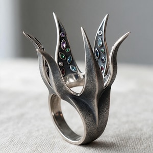 May include: A dark oxidized silver ring featuring a tall, multi-pronged structure resembling a crown or flower. The inner surfaces of the prongs are inlaid with small, multi-colored marquise and round-cut gemstones in shades of blue, green, and red.