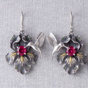 May include: A pair of silver drop earrings featuring a dark oxidised flower with a central round red gemstone, accented by small green and purple stones. A silver hummingbird is positioned on each flower, facing the central red gemstone.